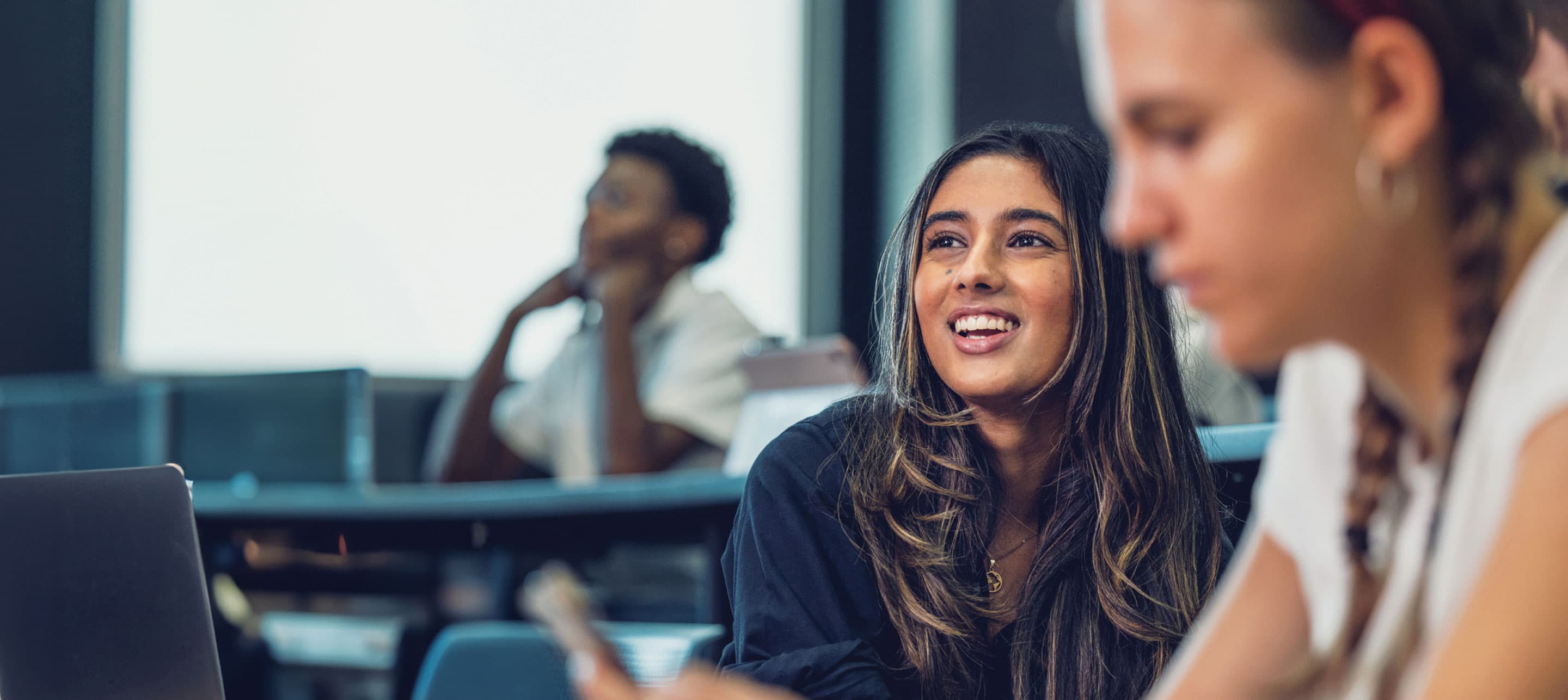 a female student in classroom in focus, with two other students in the foreground and background