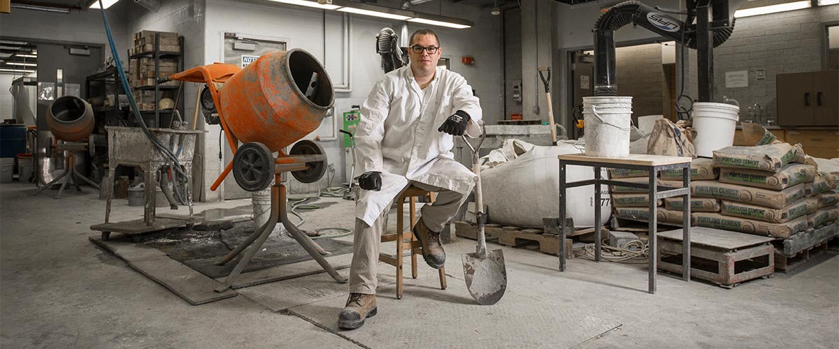 Civil Engineering graduate student with shovel, next to cement mixer in engineering building lab