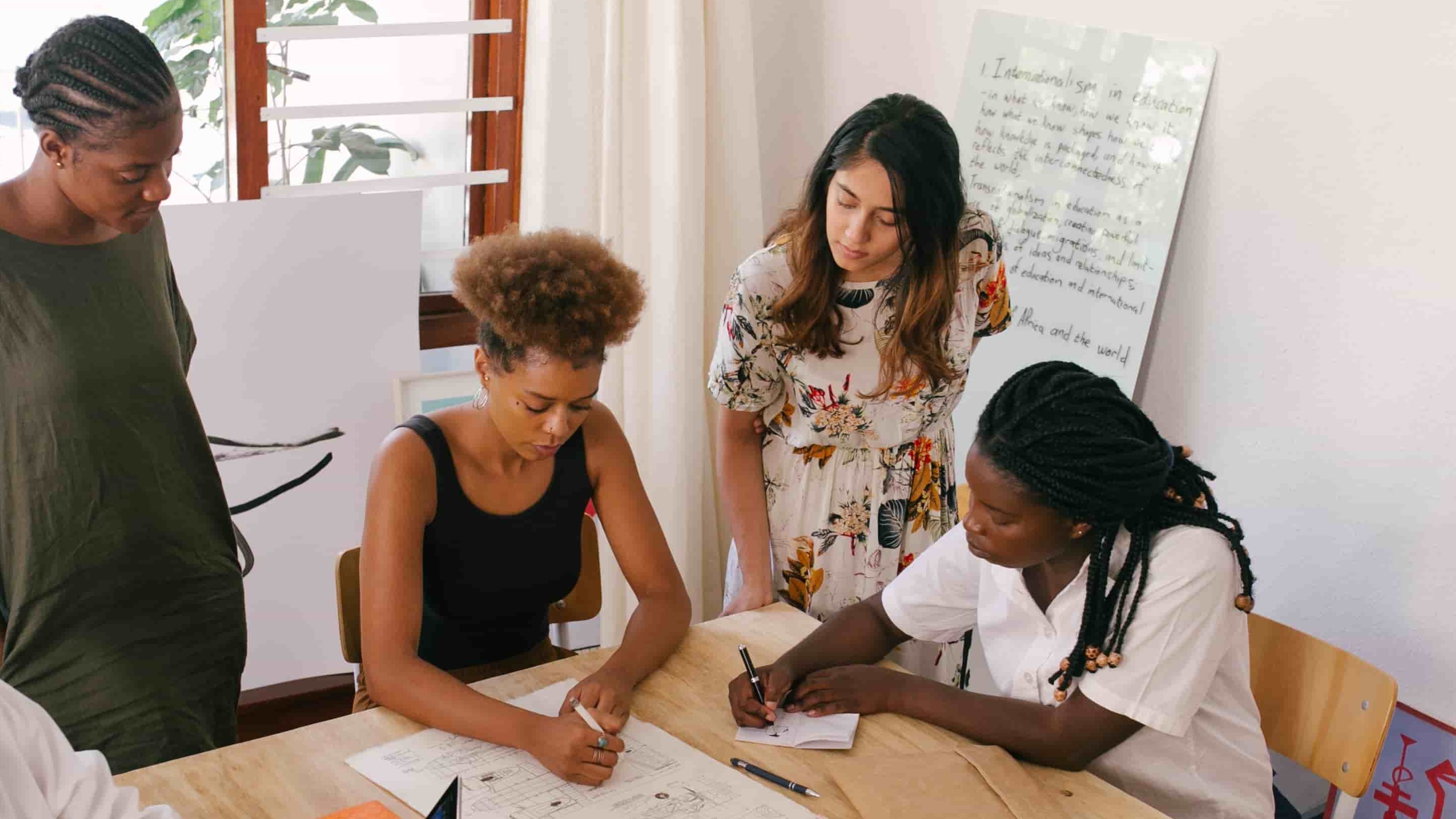 Four people around a table are collaborating and looking at a colleague's writing in a notebook.