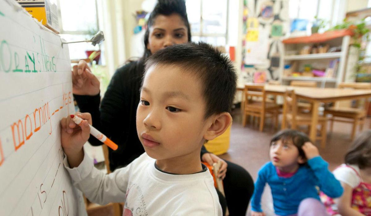 Child writing on a whiteboard with teacher and children behind sat on the floor