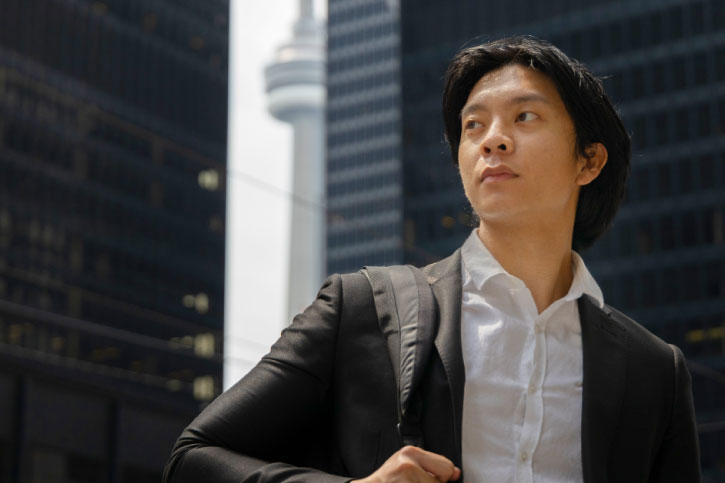 A close up of a student in the financial district holding a backpack and looking away from the camera. The CN tower is in the background.