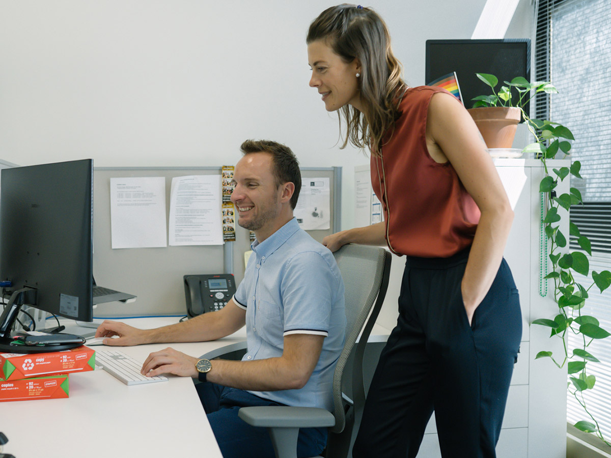 Two members of the RO team smile as they review content on a desktop computer. In the background, a Pride flag is displayed along with a thriving office plant.