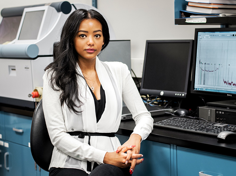 Photo of Gabriella Mankovskii, wearing a white sweater with a black belt, seated in a lab with computers behind her.