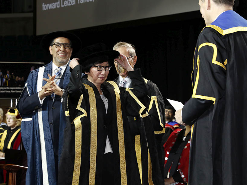 Chancellor Fukakusa on the convocation stage, while then-chancellor Lawrence Bloomberg places a cap on her head at her installation ceremony. The chancellor is surrounded by the TMU president and university administrators. 