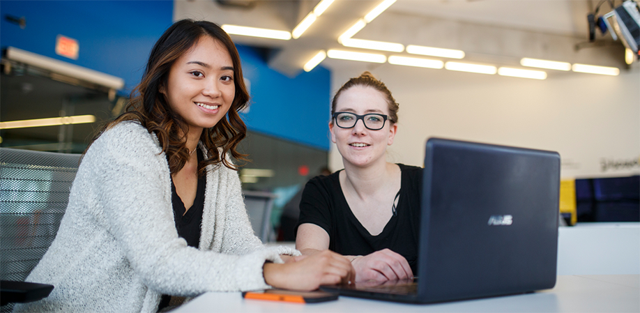 Two female students studying together at the Digital Media Experience Lab in the SLC. They are looking at the camera.